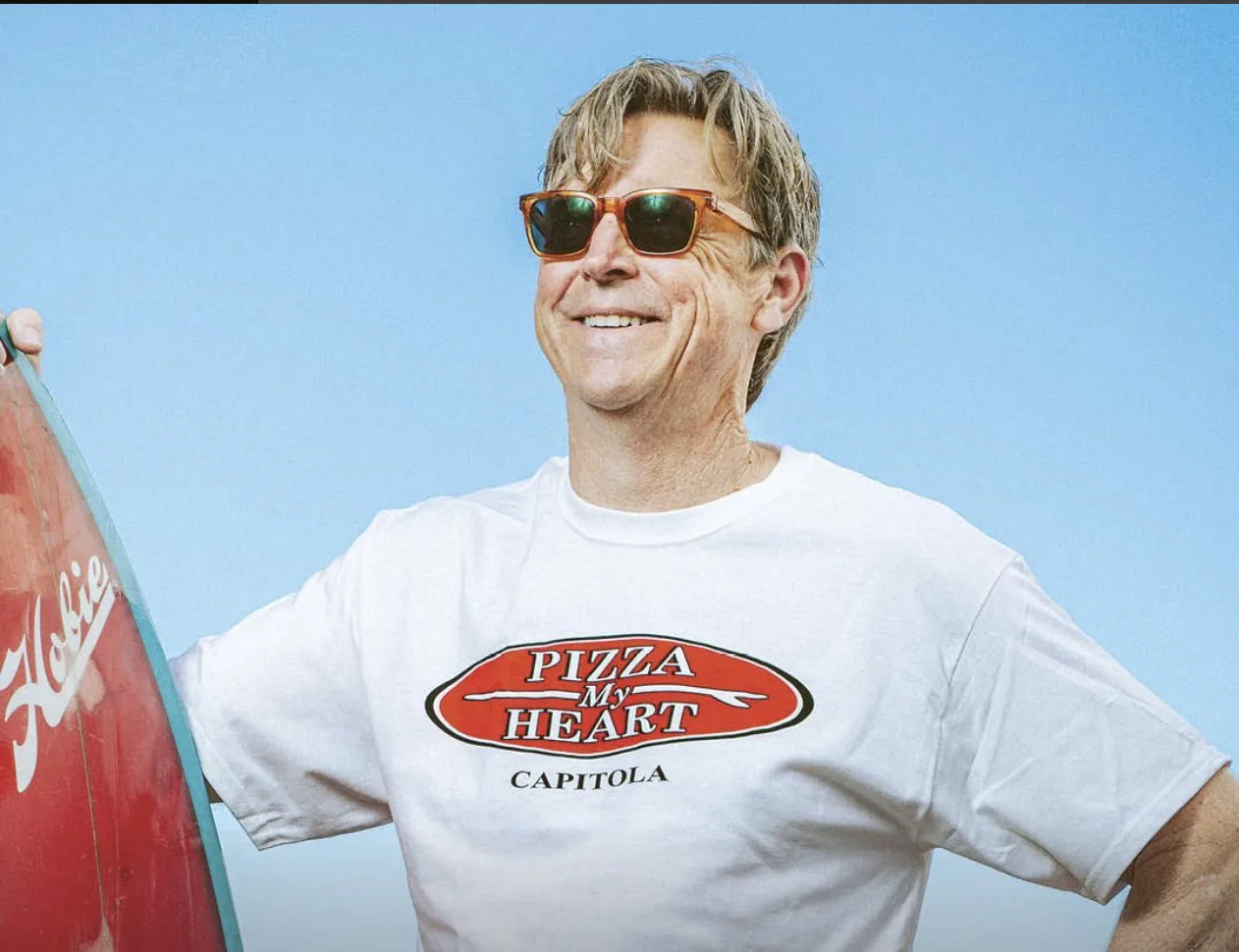 Man wearing Pizza My Heart Capitola t-shirt holding surfboard against blue sky, representing food service brand identity.