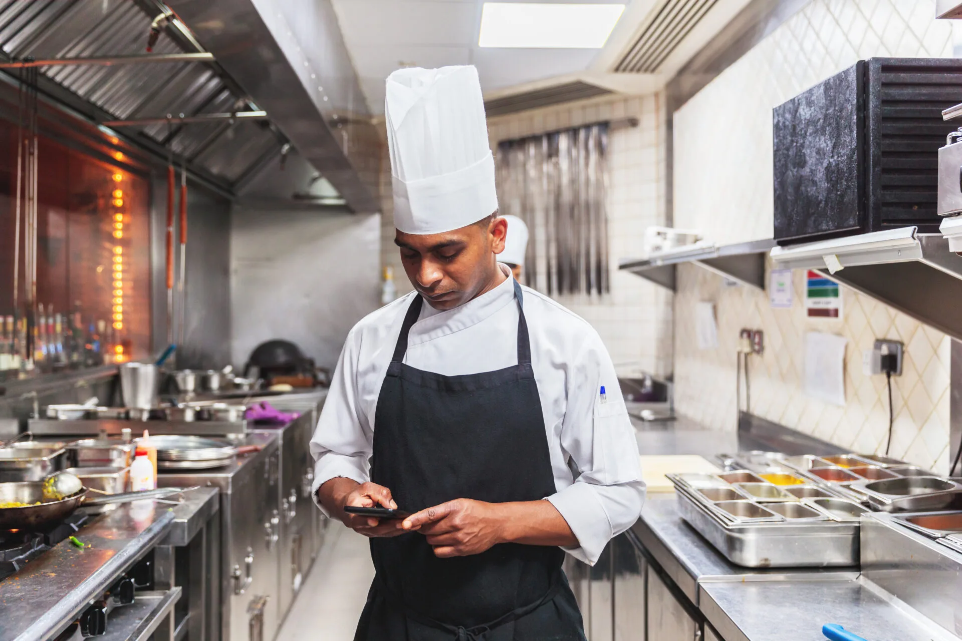Professional chef in white chef's coat and black apron uses tablet in modern commercial kitchen with stainless steel equipment and prep stations.
