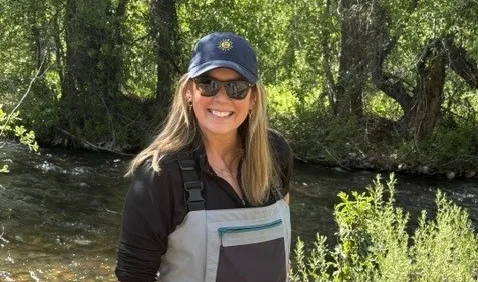 Smiling woman in a navy cap and waders standing beside a sunlit river with green trees in the background.