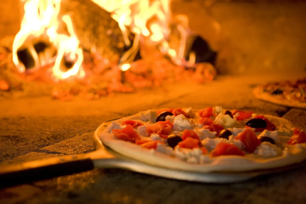 Fresh pizza with red peppers and olives on wooden peel in front of blazing wood-fired oven, ready for commercial food service.